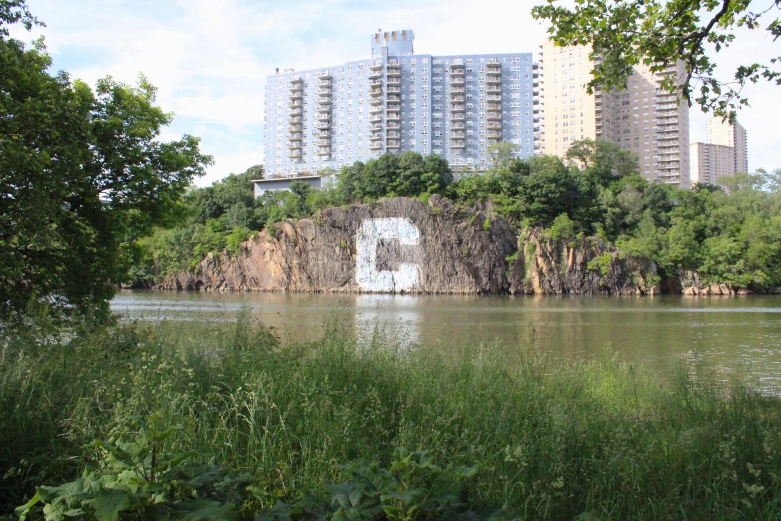 Inwood Hill Park Looking across a calm inlet toward dramatic basalt rock cliffs on the Bronx side, with a large letter "C" painted on the rock face and high-rise apartment buildings looming above.