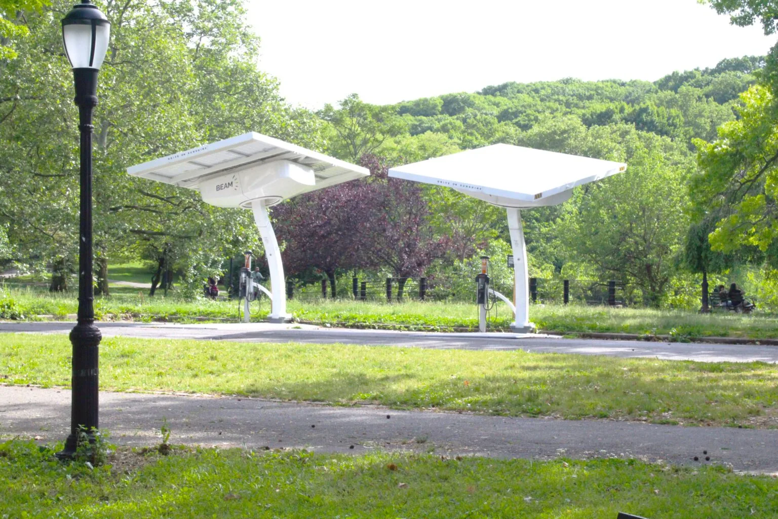 Inwood Hill Park Two solar-powered charging stations (branded "Beam") stand on a grassy lawn beside a park path, with a traditional black park lamp post nearby and forested hills in the background.