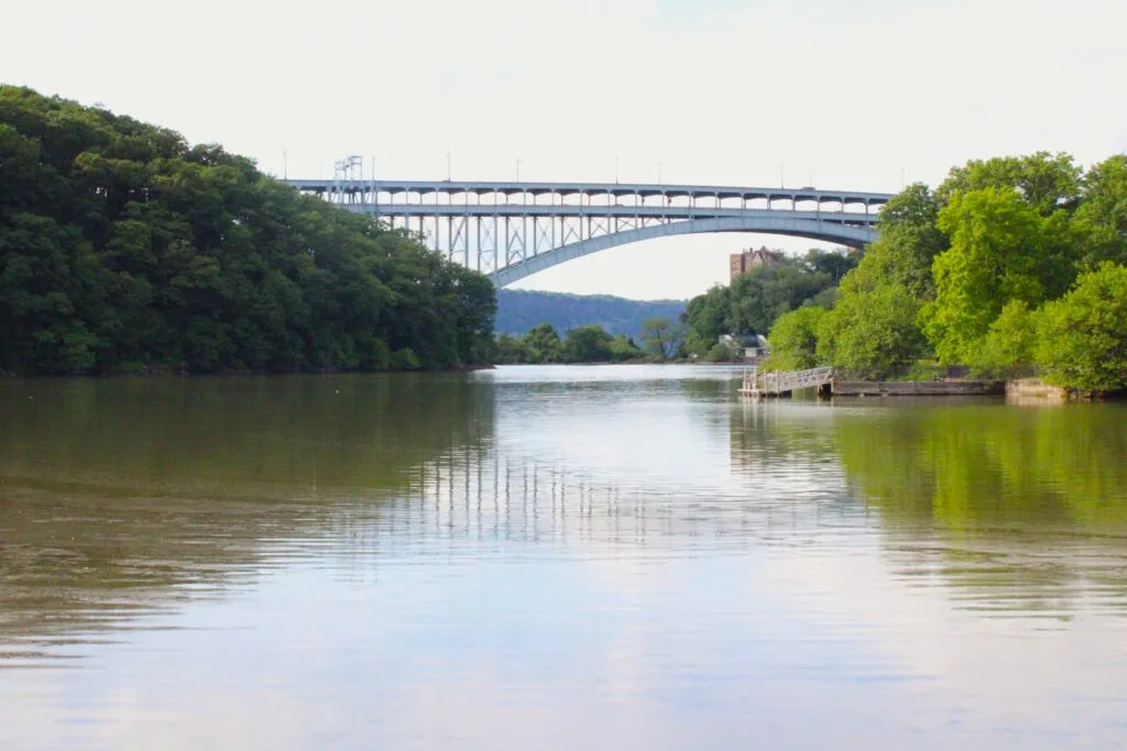 A wide view of the Harlem River Ship Canal with the Henry Hudson Bridge arching gracefully in the background, surrounded by forested hillsides and a small dock on the right bank.