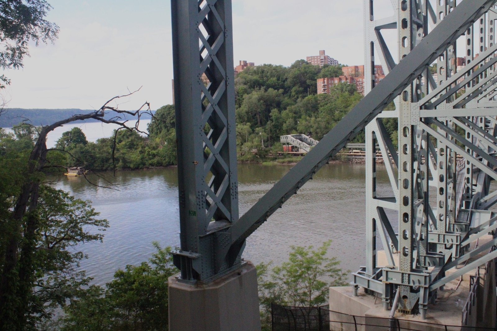 Inwood Hill Park A close-up view of one of the Henry Hudson Bridge's massive grey steel support columns and diagonal truss members, with the Harlem River and wooded Bronx shoreline visible beyond.