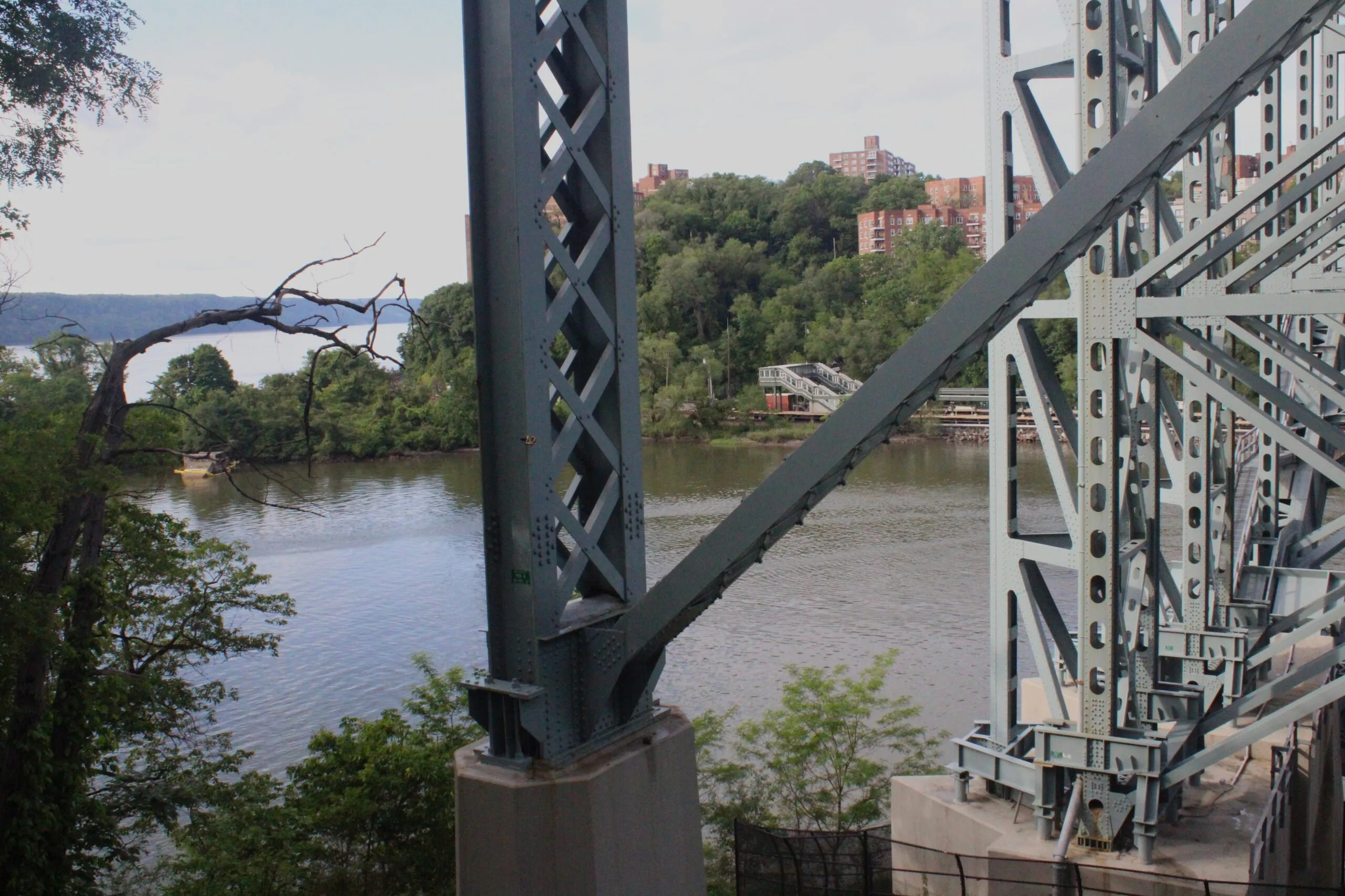 Inwood Hill Park A close-up view of one of the Henry Hudson Bridge's massive grey steel support columns and diagonal truss members, with the Harlem River and wooded Bronx shoreline visible beyond.